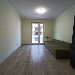 ExpressRemont - Wnętrze pokoju po remoncie z panelami podłogowymi imitującymi drewno, świeżo pomalowanymi ścianami na jasnozielony kolor, z widokiem na budynek w budowie przez balkonowe drzwi i z częściowo...