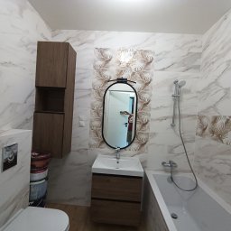 ExpressRemont - Nowoczesna łazienka z marmurowymi płytkami, szafką wiszącą z drewnianym frontem, owalnym lustrem z podświetleniem i widocznymi pojemnikami z farbami oraz silikonami.