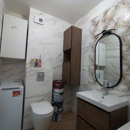 ExpressRemont - Wykończona łazienka z pralką, umywalką z szafką w kolorze drewna, toaletą, wysoką szafką i stosem puszek po farbie przy ścianie z marmurkowymi i liściastymi wzorami na płytkach.