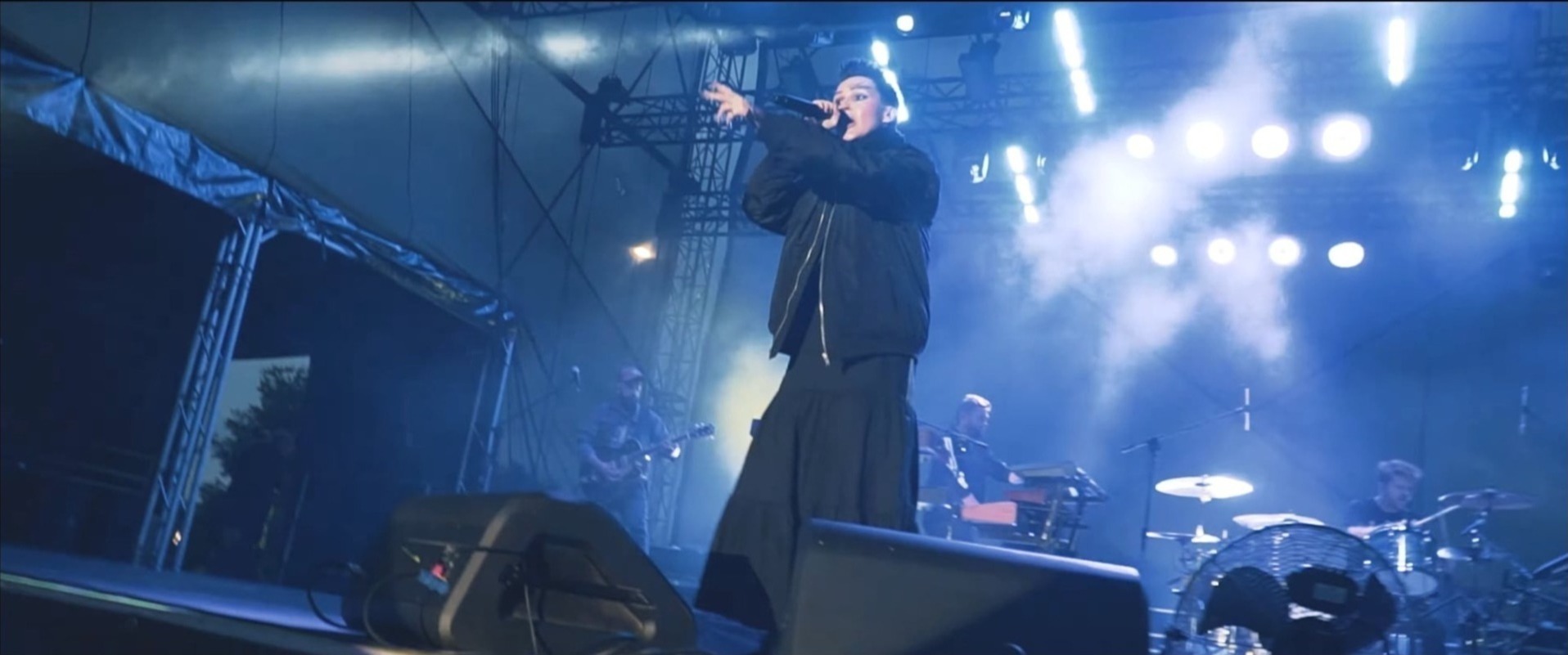 Wokalistka w czarnym stroju na scenie podczas koncertu, oświetlona niebieskim światłem, z zespołem w tle. Ujęcie z dołu, widoczna konstrukcja sceny.
