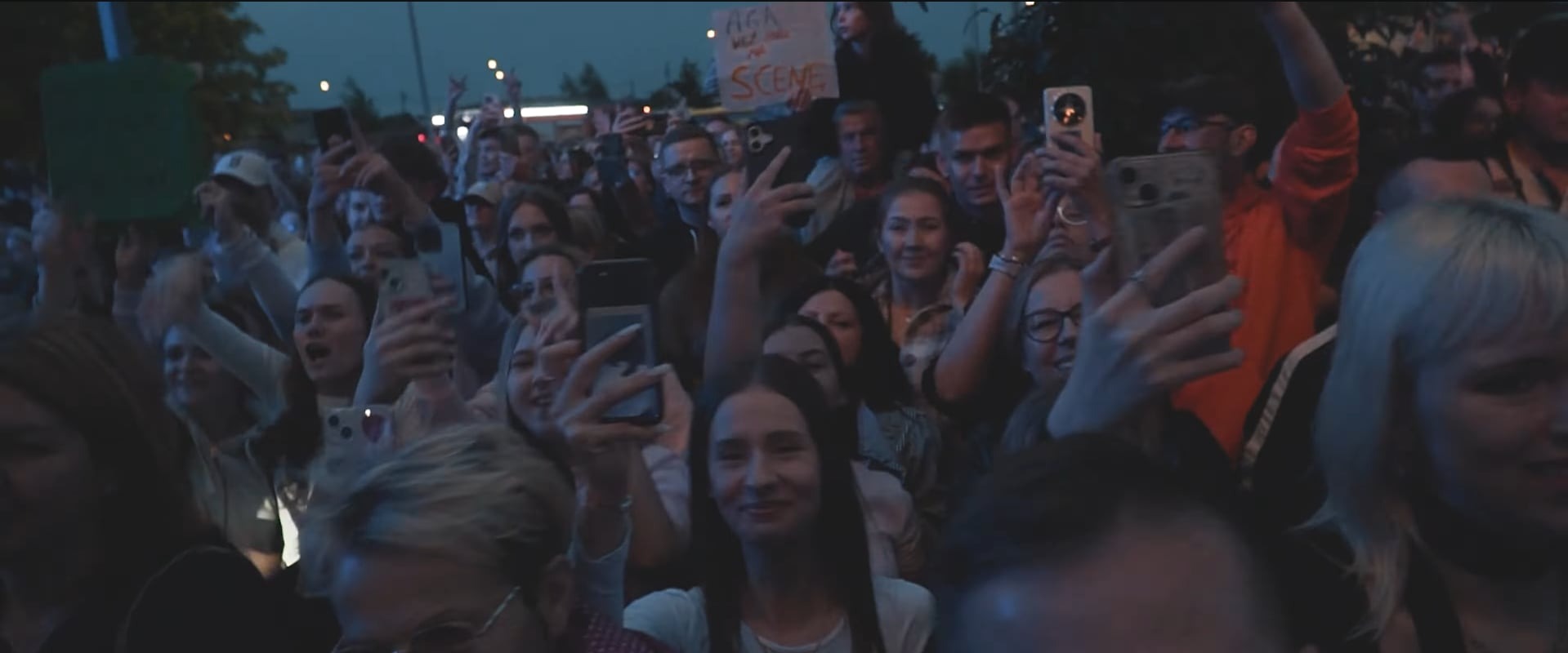 Tłum ludzi z podniesionymi telefonami podczas koncertu. W tle widoczny napis 'AGA WEZ MNIE NA SCENE'. Ujęcie z dołu, ciemne oświetlenie.