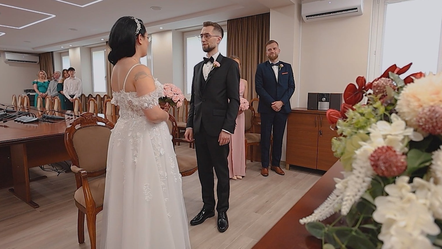 Sala ślubów: panna młoda w białej sukni, pan młody w czarnym garniturze, goście w tle. Ujęcie z boku, kwiaty na pierwszym planie, jasne wnętrze.