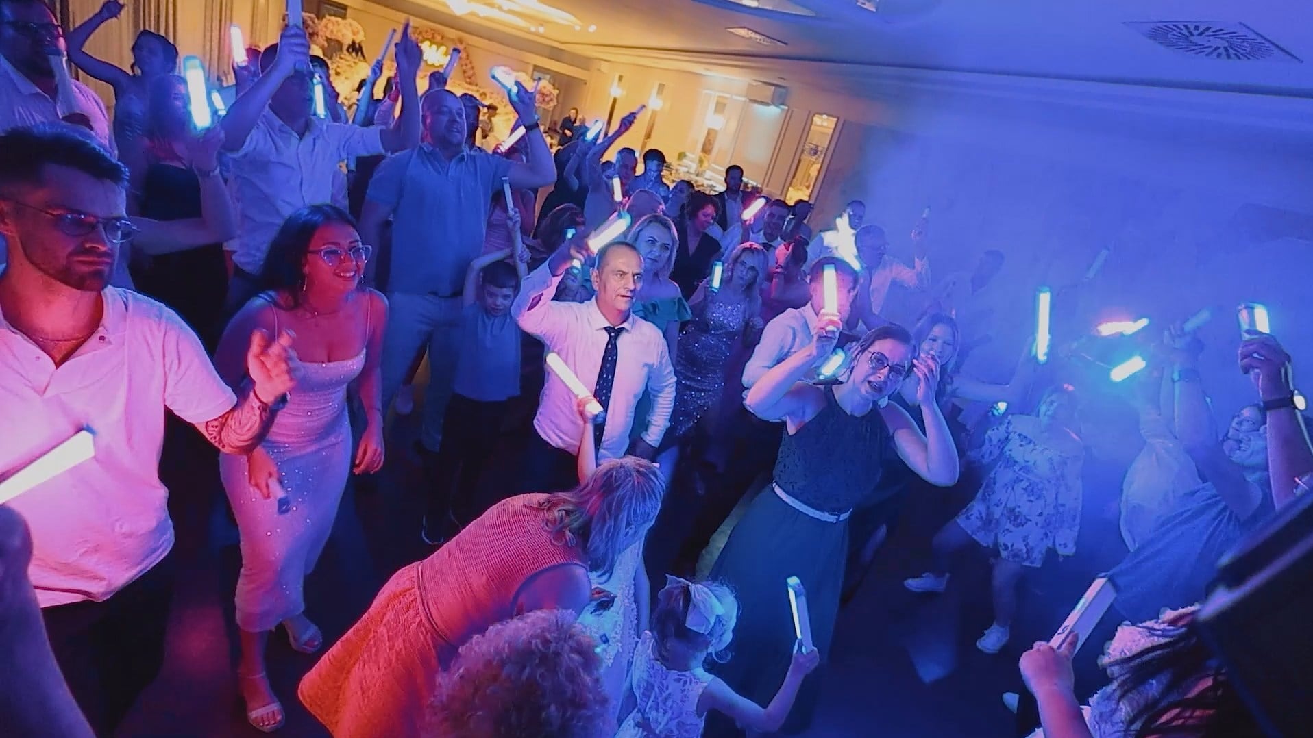 Roztańczony tłum gości weselnych w niebieskim oświetleniu, trzymających świecące pałeczki. Dynamiczne ujęcie z perspektywy żabiej.