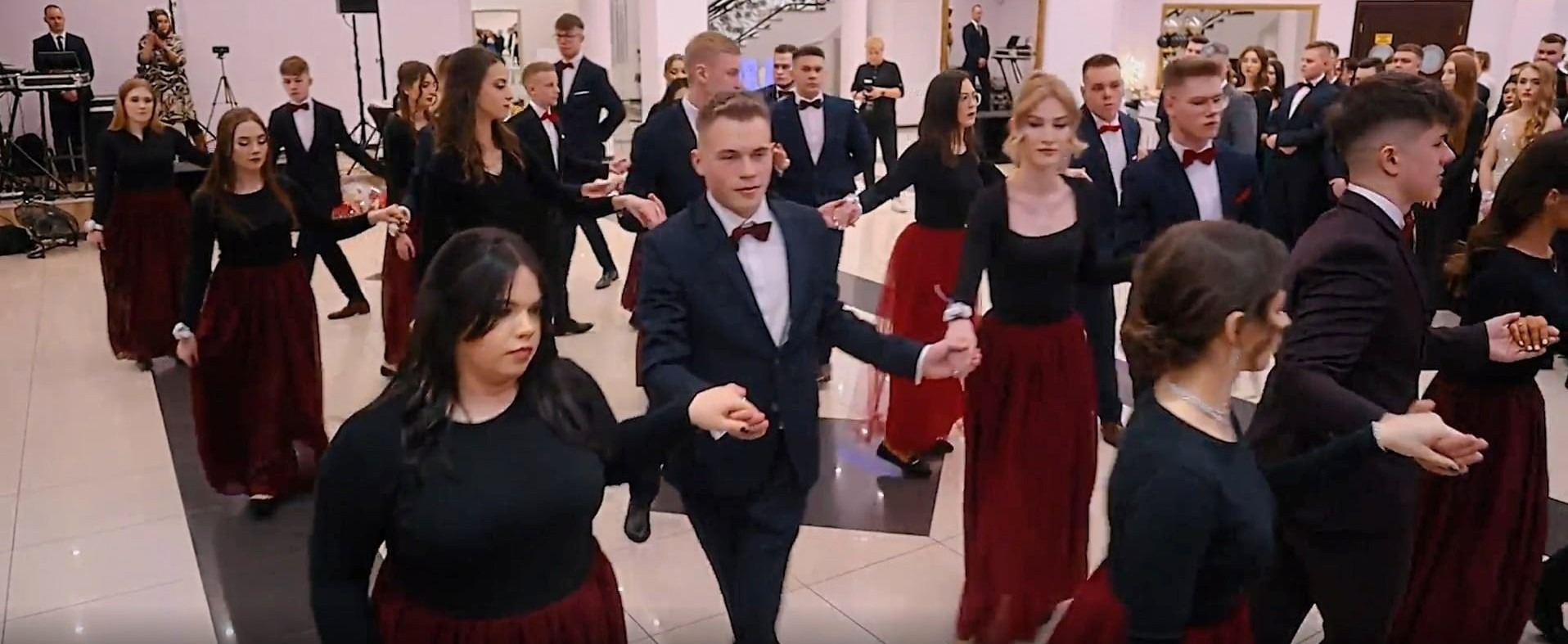 Uczestnicy studniówki w trakcie poloneza. Młodzież w strojach wieczorowych, dziewczęta w bordowych spódnicach i czarnych bluzkach, chłopcy w garniturach z muszkami.