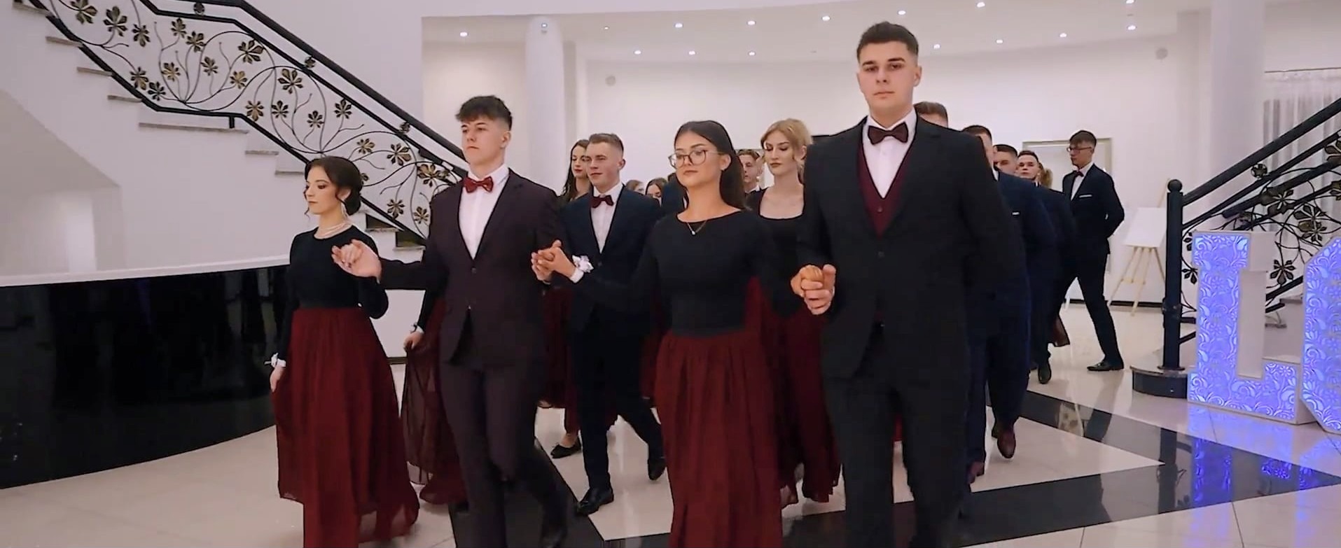 Polonez studniówkowy: Młodzież w strojach wieczorowych tańczy w eleganckiej sali balowej z dekoracyjnymi schodami i podświetlanymi literami. Ujęcie z kamery podczas nagrania.