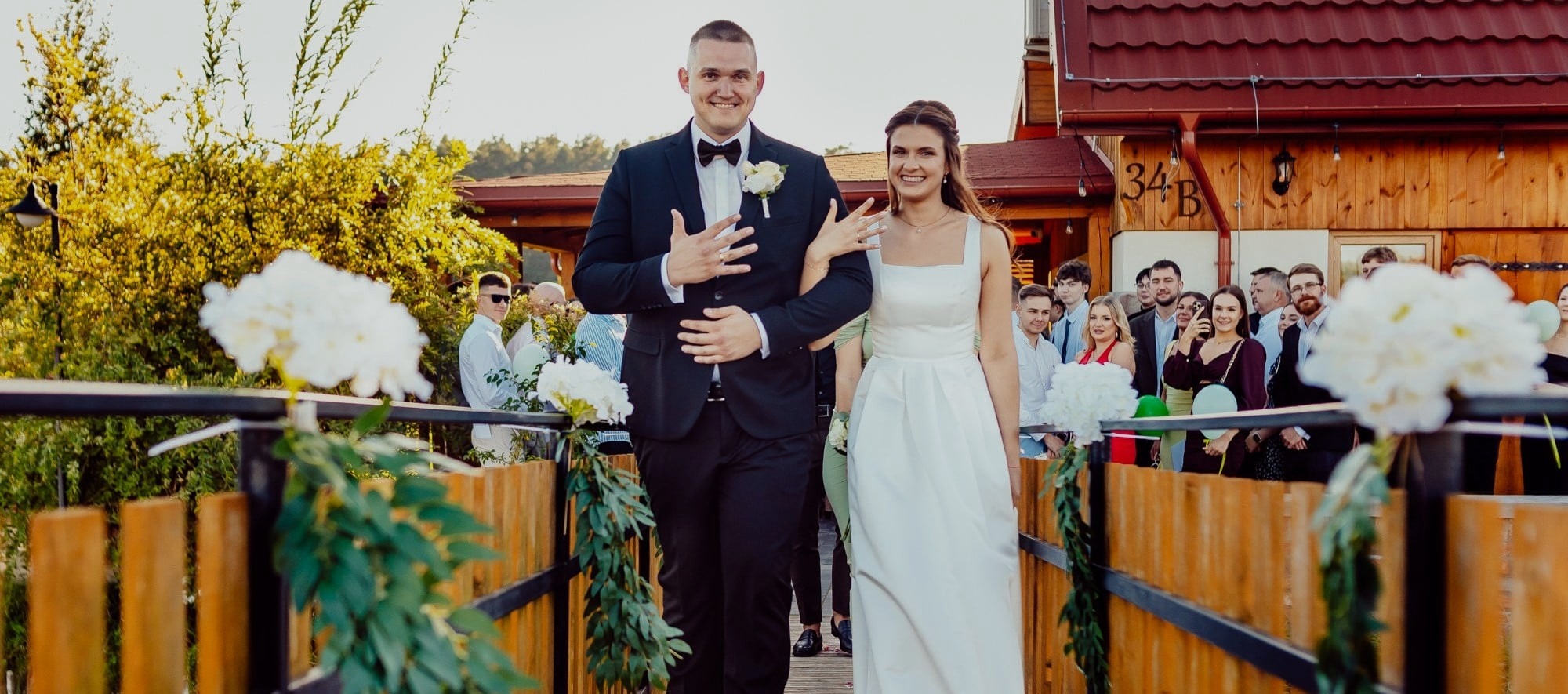 Uśmiechnięta para młoda, on w czarnym smokingu, ona w białej sukni, idą udekorowanym mostem, w tle goście weselni i budynek z numerem 34B.