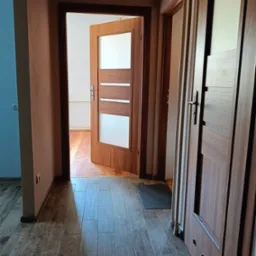 Wnętrze przedpokoju z widocznymi drzwiami wewnętrznymi, panelami podłogowymi w odcieniach brązu i beżu, oraz fragmentem pomalowanej na niebiesko ściany.