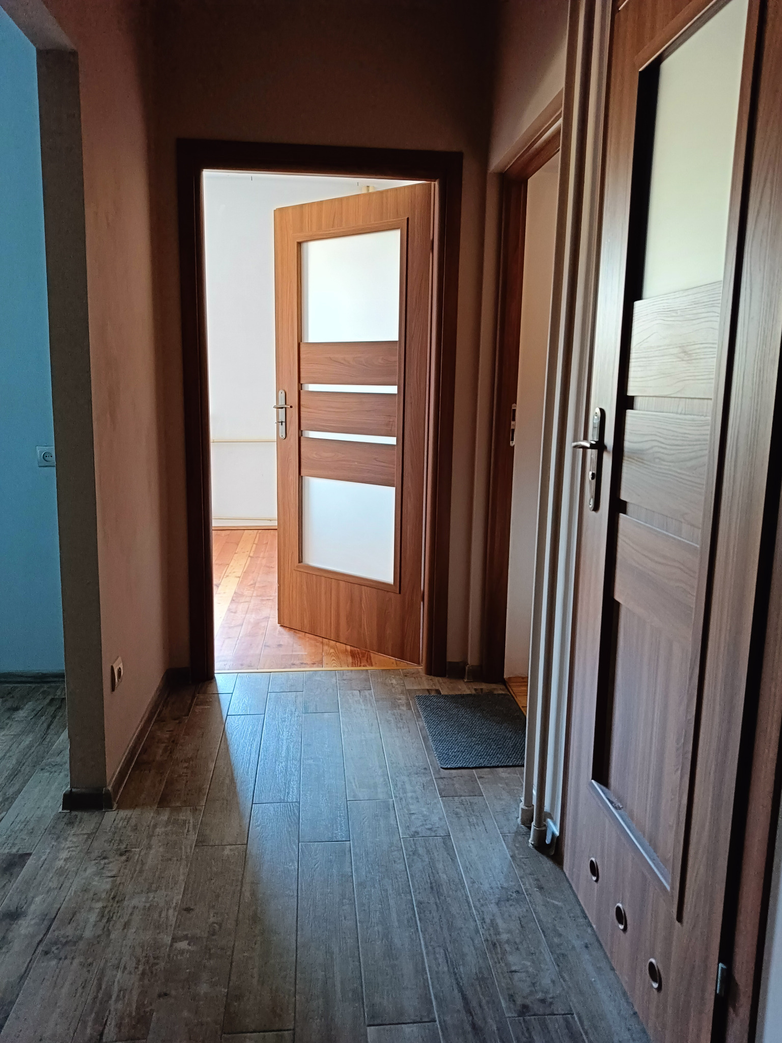 Wnętrze przedpokoju z widocznymi drzwiami wewnętrznymi, panelami podłogowymi w odcieniach brązu i beżu, oraz fragmentem pomalowanej na niebiesko ściany.