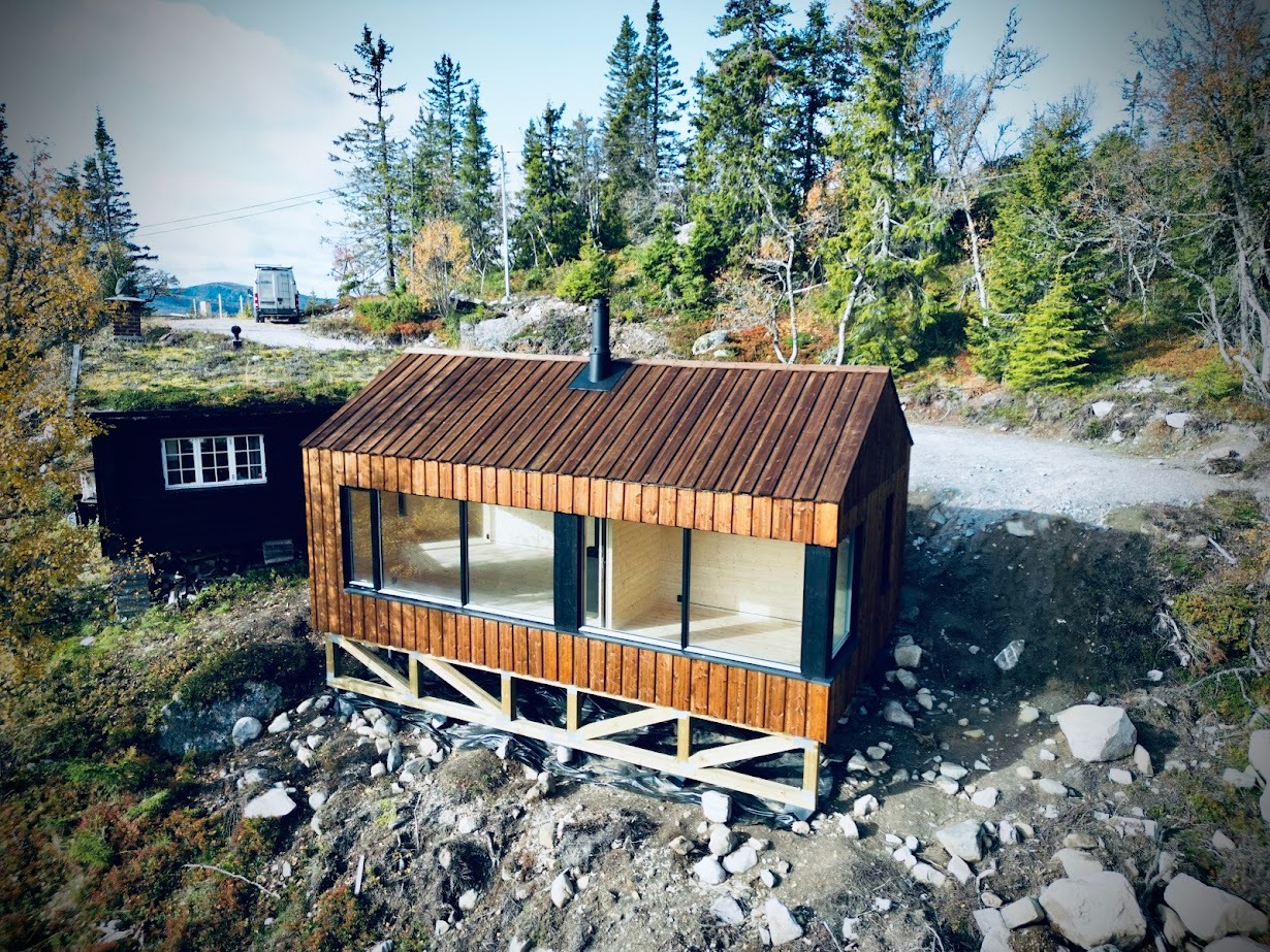 Nowoczesny, drewniany domek letniskowy z dużymi oknami, posadowiony na fundamencie palowym, obok starszego budynku z zielonym dachem, w otoczeniu drzew i skał.