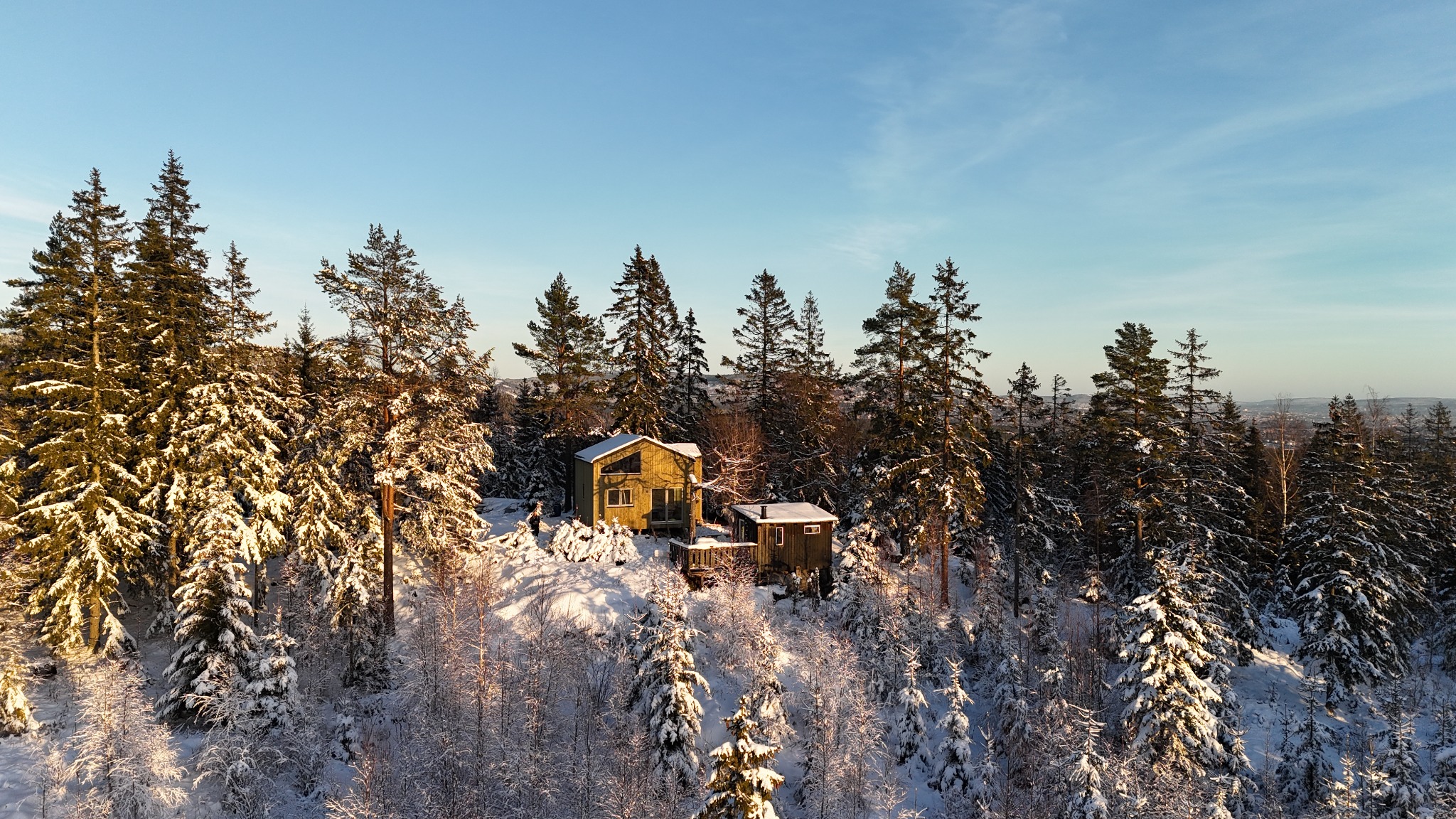 Dwa drewniane domki szkieletowe w zimowym lesie, jeden z oknem trójkątnym na szczycie, oba z widocznymi kominami, pokryte śniegiem.