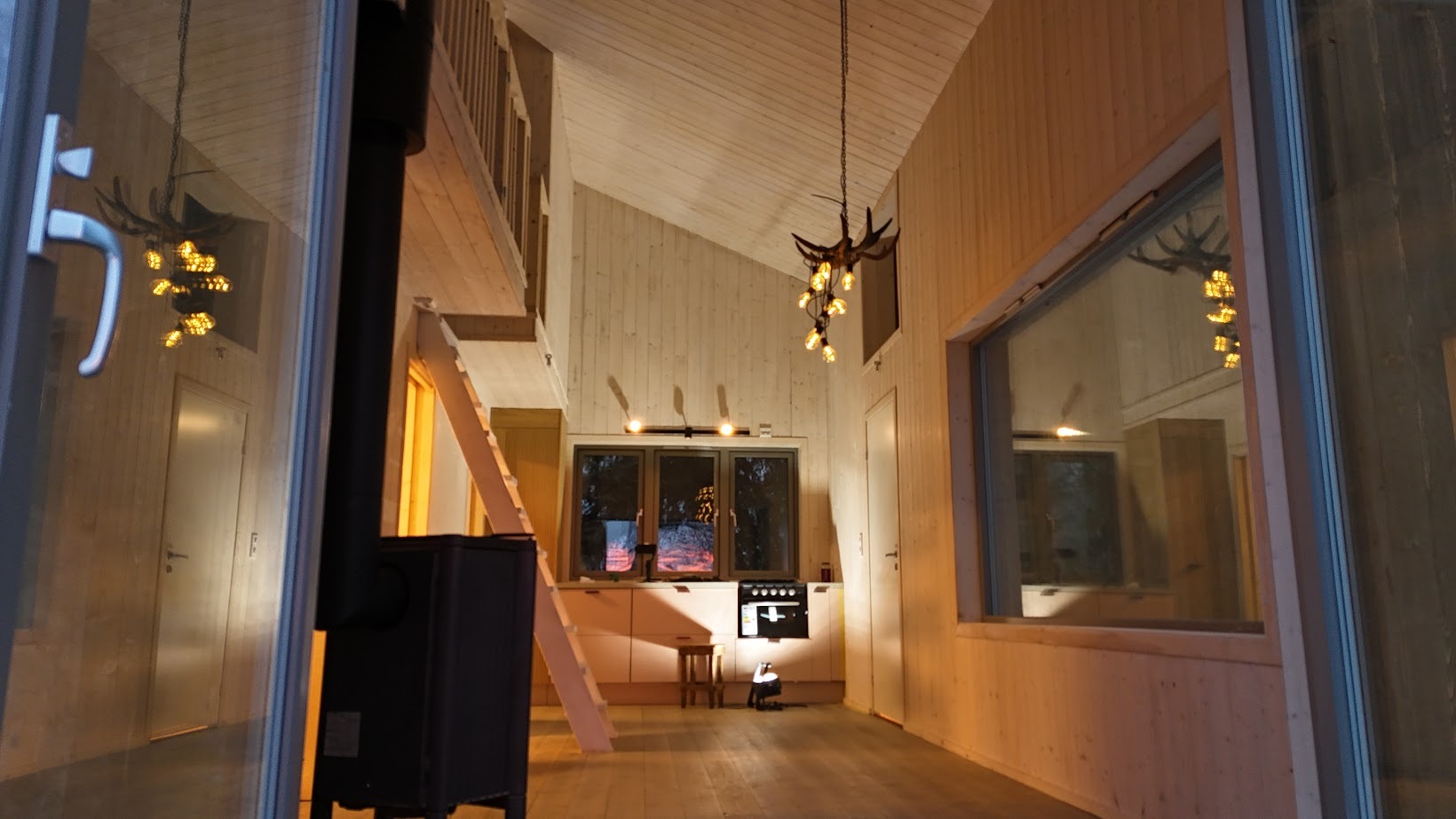 Wnętrze domu z drewnianą boazerią, widok na kuchnię z oknami, kominek i schody na antresolę, oświetlone lampą w kształcie poroża.