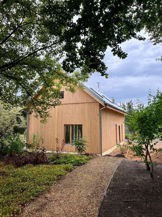 Nowoczesny dom szkieletowy z jasnego drewna z panelami słonecznymi na dachu, otoczony zielenią i ścieżką z kamyków.