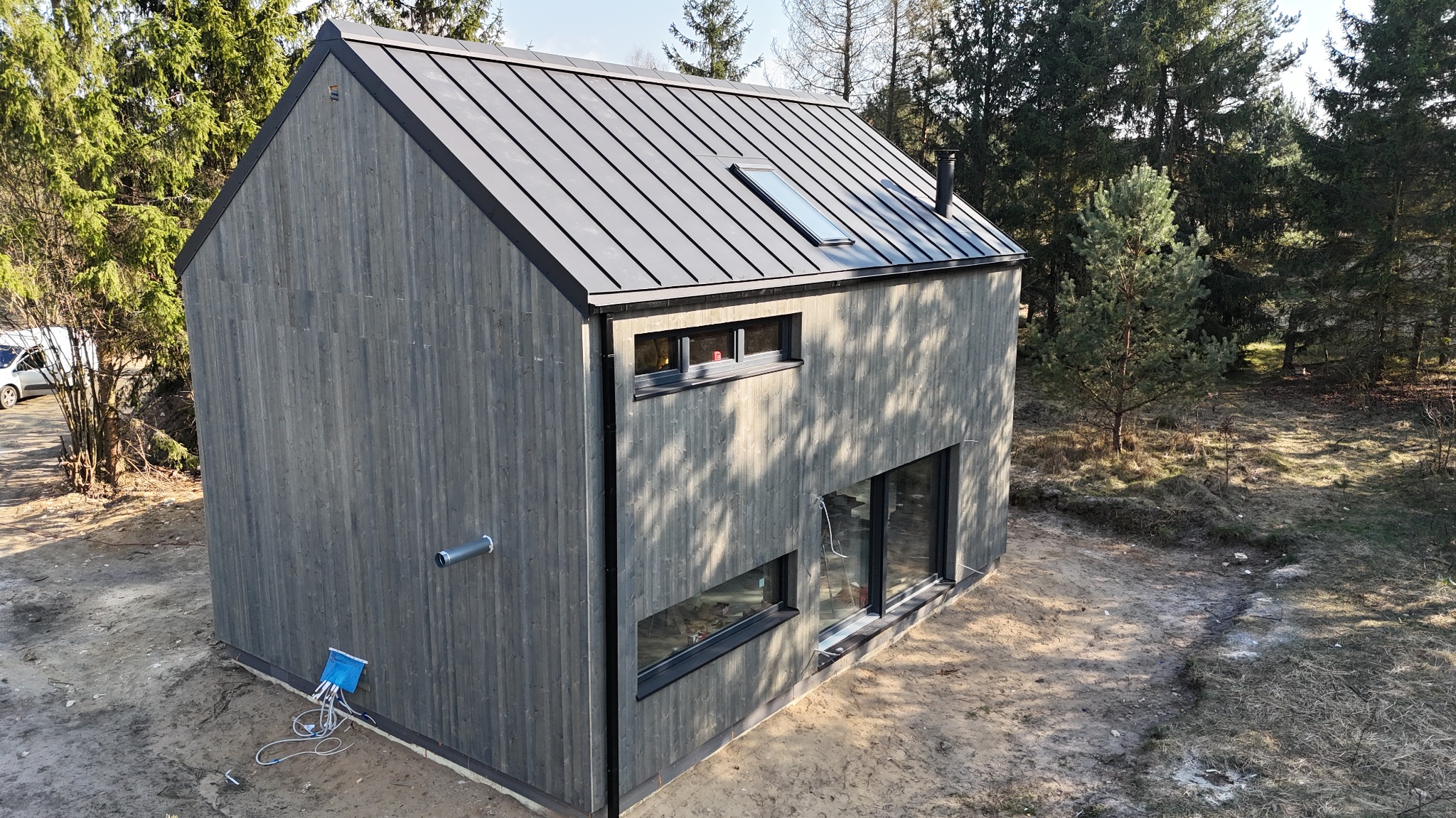 Nowoczesny, dwukondygnacyjny dom z szarej deski z metalowym dachem i oknami dachowymi w Częstochowie, otoczony drzewami, widoczne przyłącza instalacji.