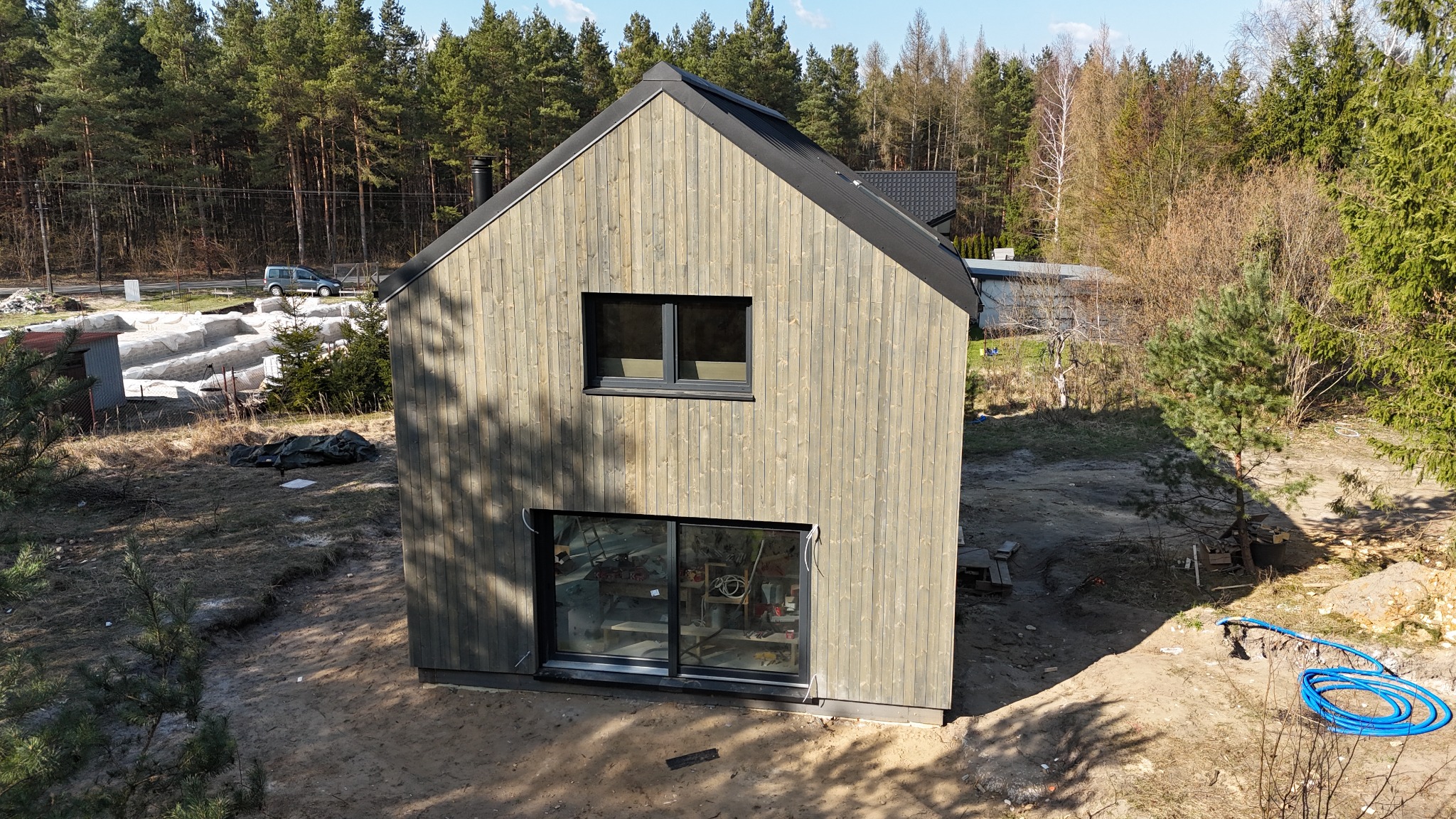 Surowy, dwukondygnacyjny budynek z szarego drewna z czarnym dachem i oknami, widok z zewnątrz na plac budowy w otoczeniu drzew.