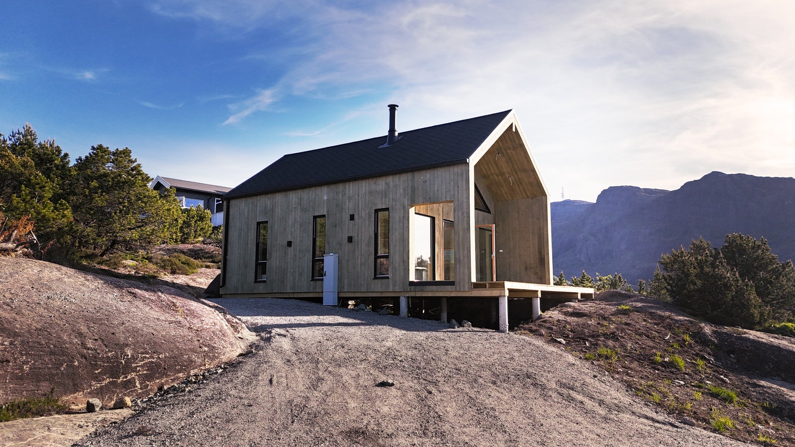 Nowoczesny dom o prostej bryle z drewnianą elewacją, czarnym dachem i kominem, posadowiony na skalistym wzgórzu z widokiem na góry w tle.