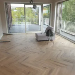 Jasne panele ułożone w jodełkę w przestronnym pokoju z wyjściem na balkon. Widoczne kartony z panelami i podkładki. Minimalistyczny design wnętrza z szarą sofą.