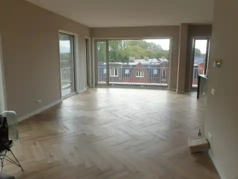 Wykończone wnętrze z parkietem w jodełkę, dużymi oknami balkonowymi i ścianami w odcieniach beżu. Widok na zabudowę miejską zza balkonu.