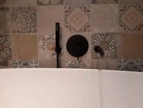 Retro łazienka: biała wanna z czarną armaturą na tle patchworkowych płytek w odcieniach beżu i szarości. Widoczny panel baterii i słuchawka prysznicowa.