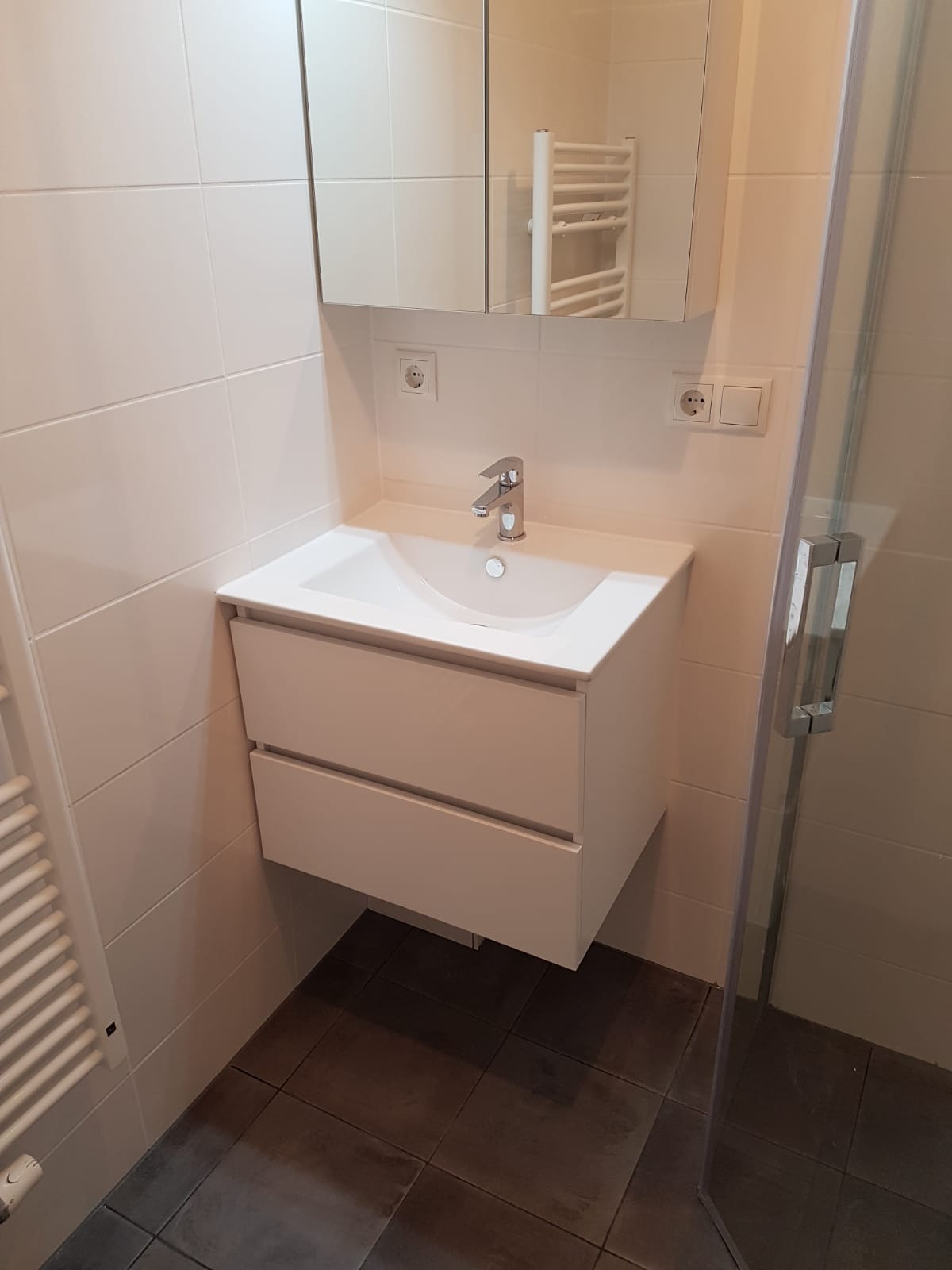 Biała umywalka z szafką i lustrem w nowoczesnej łazience z beżowymi płytkami i ciemną podłogą. Widoczny fragment kabiny prysznicowej i grzejnika.
