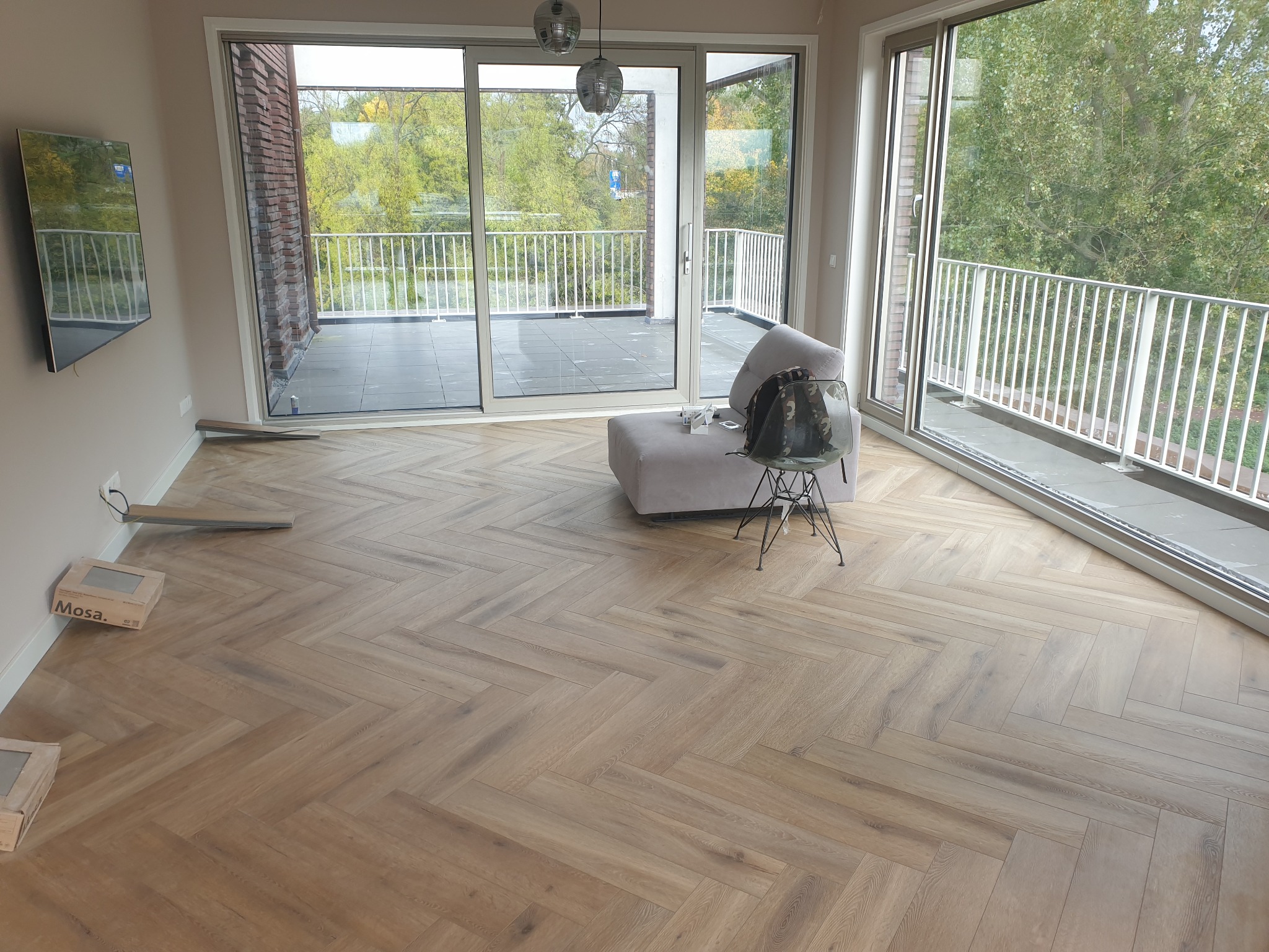 Jasne panele ułożone w jodełkę w przestronnym pokoju z wyjściem na balkon. Widoczne kartony z panelami i podkładki. Minimalistyczny design wnętrza z szarą sofą.