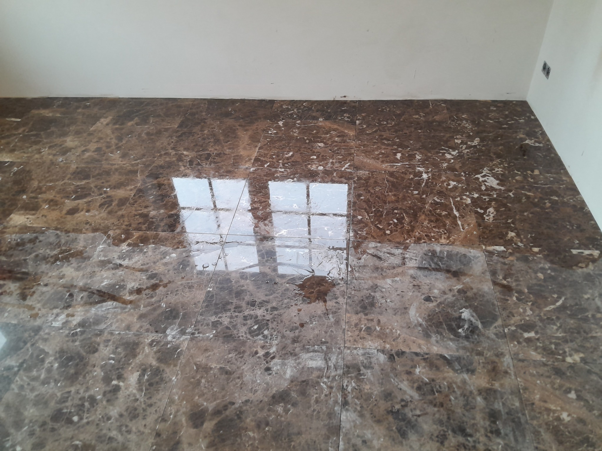 Błyszcząca, brązowa marmurowa podłoga po ułożeniu płytek, z widocznym odbiciem światła okiennego i gniazdkiem elektrycznym przy ścianie.