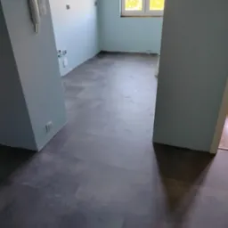 Nowa, ciemnoszara podłoga z paneli w jasnym, odświeżonym pokoju z niebieskimi ścianami, widoczne gniazdka elektryczne i okno z widokiem na drzewa.