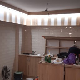 Tomasz Lewandowski - Wnętrze kuchni w trakcie prac wykończeniowych: ułożone płytki na ścianie, zamontowane szafki z jasnego drewna z widocznym zlewem, puszki po piwie, wiadro z farbą i narzędzia malarskie na podłodze...