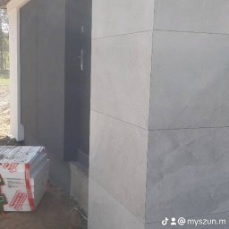 Tomasz Lewandowski - Wykończona elewacja domu z szarych płyt imitujących kamień, widoczny fragment drzwi wejściowych i stos materiałów budowlanych przed budynkiem.
