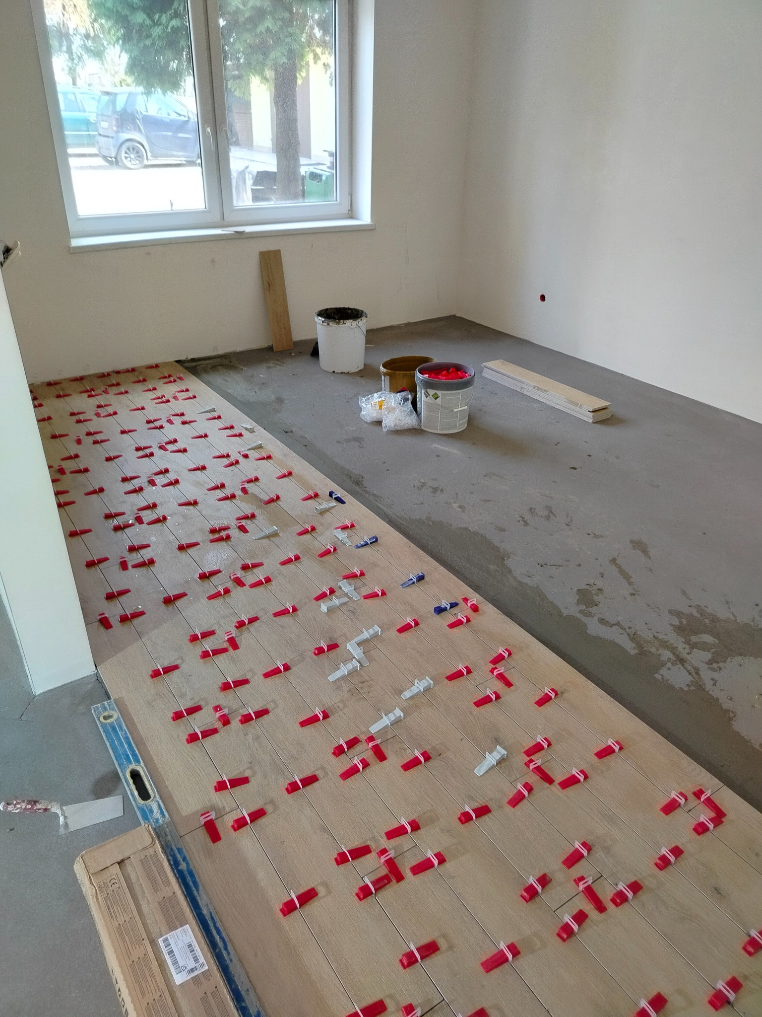 Układanie podłogi z paneli drewnopodobnych z użyciem systemu poziomowania; widoczne kliny i poziomica, w tle puste ściany i okno.