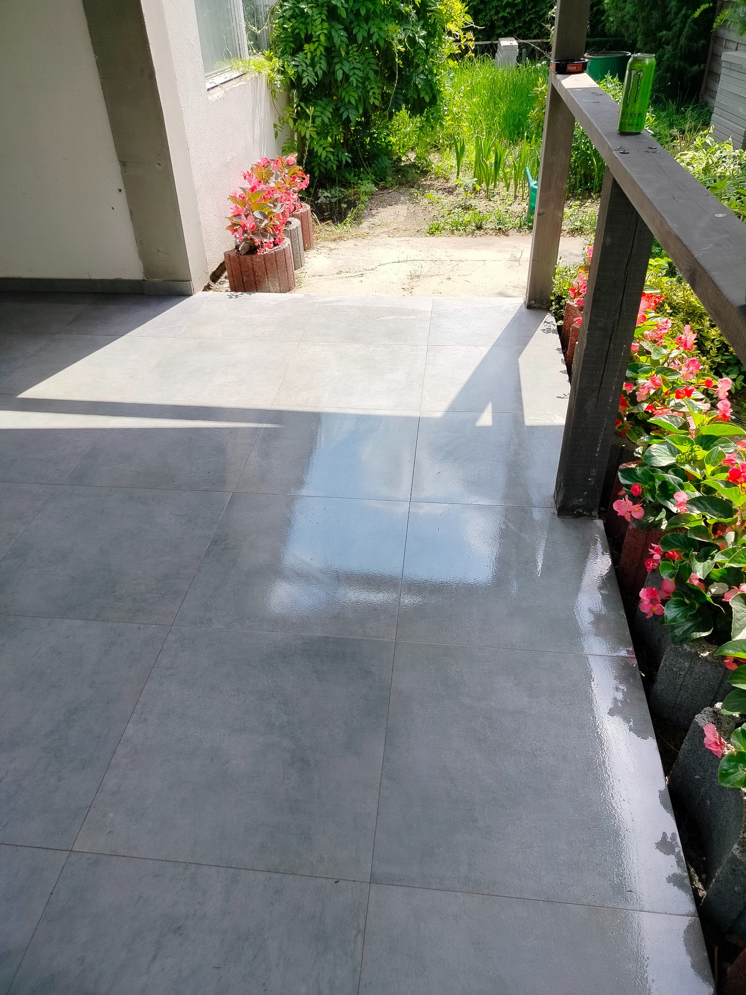 Świeżo ułożona, błyszcząca, szara terakota na zewnątrz, obok drewnianej balustrady z kwiatami w doniczkach, odbijająca światło słoneczne.