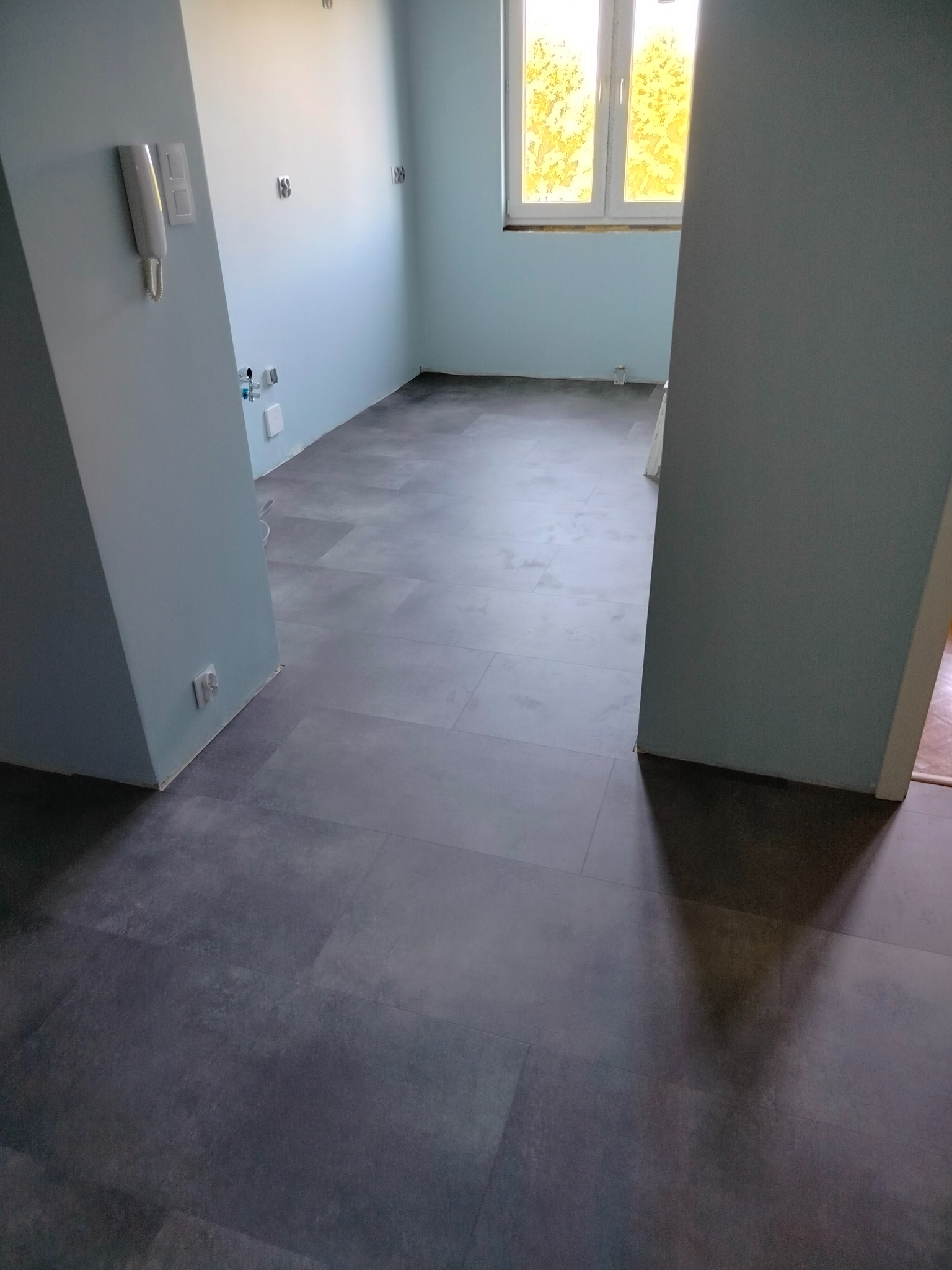 Nowa, ciemnoszara podłoga z paneli w jasnym, odświeżonym pokoju z niebieskimi ścianami, widoczne gniazdka elektryczne i okno z widokiem na drzewa.