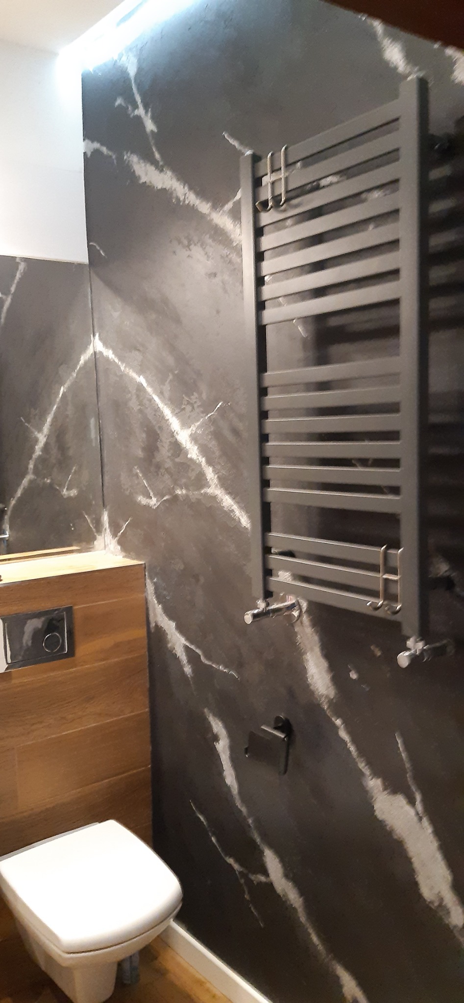 Nowoczesna łazienka z szarą ścianą imitującą marmur, grzejnikiem drabinkowym i podwieszaną toaletą.