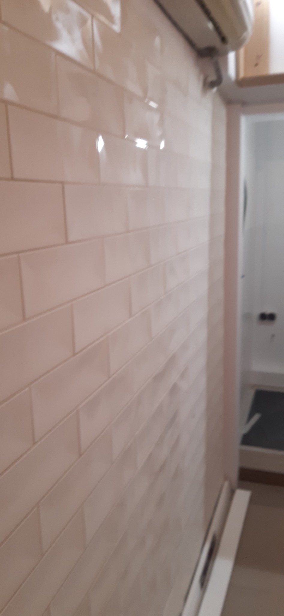 Ściana wyłożona błyszczącymi, beżowymi płytkami w łazience, widoczny fragment klimatyzatora i wejście do pomieszczenia z szarą wycieraczką.