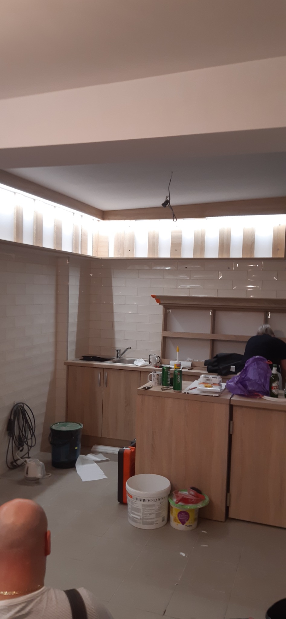 Wnętrze kuchni w trakcie prac wykończeniowych: ułożone płytki na ścianie, zamontowane szafki z jasnego drewna z widocznym zlewem, puszki po piwie, wiadro z farbą i narzędzia malarskie na podłodze...