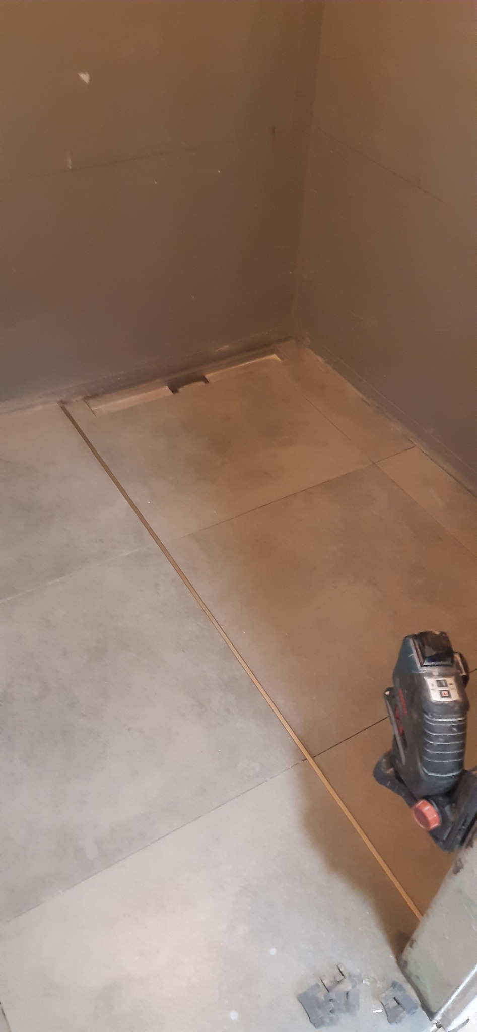 Układanie płytek podłogowych w łazience, widoczny laser krzyżowy do poziomowania, szare ściany i płytki, częściowo zakryta studzienka odpływowa.