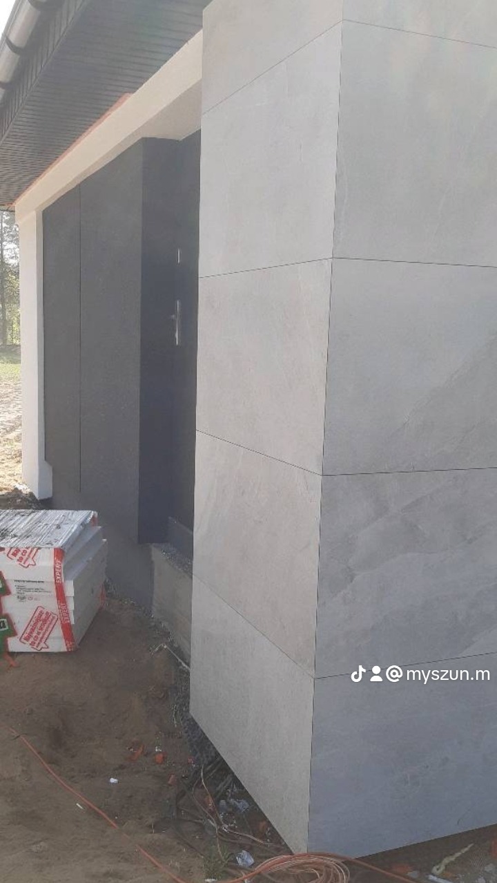Wykończona elewacja domu z szarych płyt imitujących kamień, widoczny fragment drzwi wejściowych i stos materiałów budowlanych przed budynkiem.