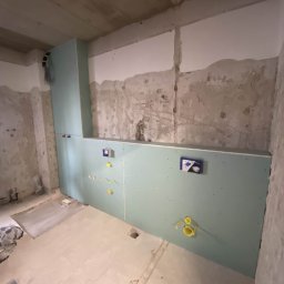 Rewall - Wnętrze łazienki w trakcie remontu: ściany z surowego betonu i fragment zabudowy z płyty karton-gips z widocznymi instalacjami wodno-kanalizacyjnymi.