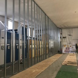 Rewall - Montaż ścianki działowej z metalowych profili w pomieszczeniu przemysłowym. Widoczne panele elektryczne i drabina. Podłoga zabezpieczona płytami OSB.
