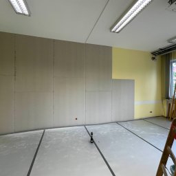 Rewall - Wnętrze w trakcie montażu płyt karton-gips na ścianie, widoczna drabina i narzędzia. Część ściany pomalowana na żółto, podłoga zabezpieczona folią.