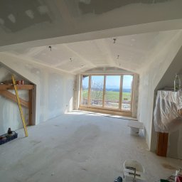 Rewall - Poddasze po szpachlowaniu i montażu płyt gipsowych, z oknem na krajobraz. Widoczne narzędzia i materiały budowlane, surowy stan wykończenia.