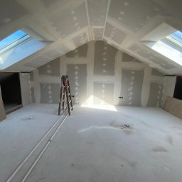 Rewall - Poddasze w trakcie wykańczania: ściany i sufit pokryte płytami gipsowo-kartonowymi, widoczne łączenia szpachlowane, drabina w centrum, okna dachowe w skosach.