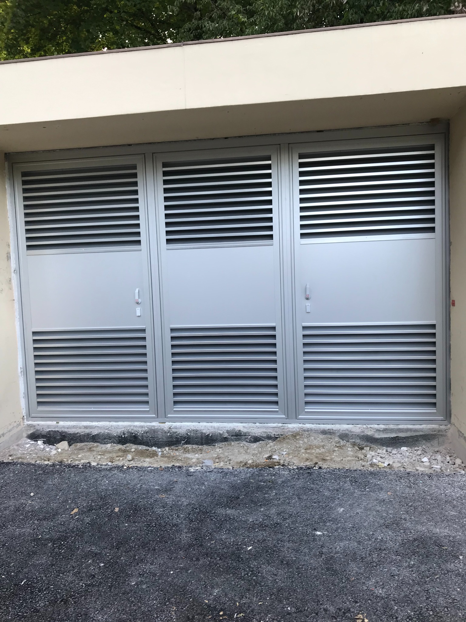Szare, metalowe, trzyskrzydłowe drzwi garażowe z poziomymi żaluzjami wentylacyjnymi, osadzone w beżowej wnęce budynku, z widocznym fragmentem asfaltowego podjazdu.