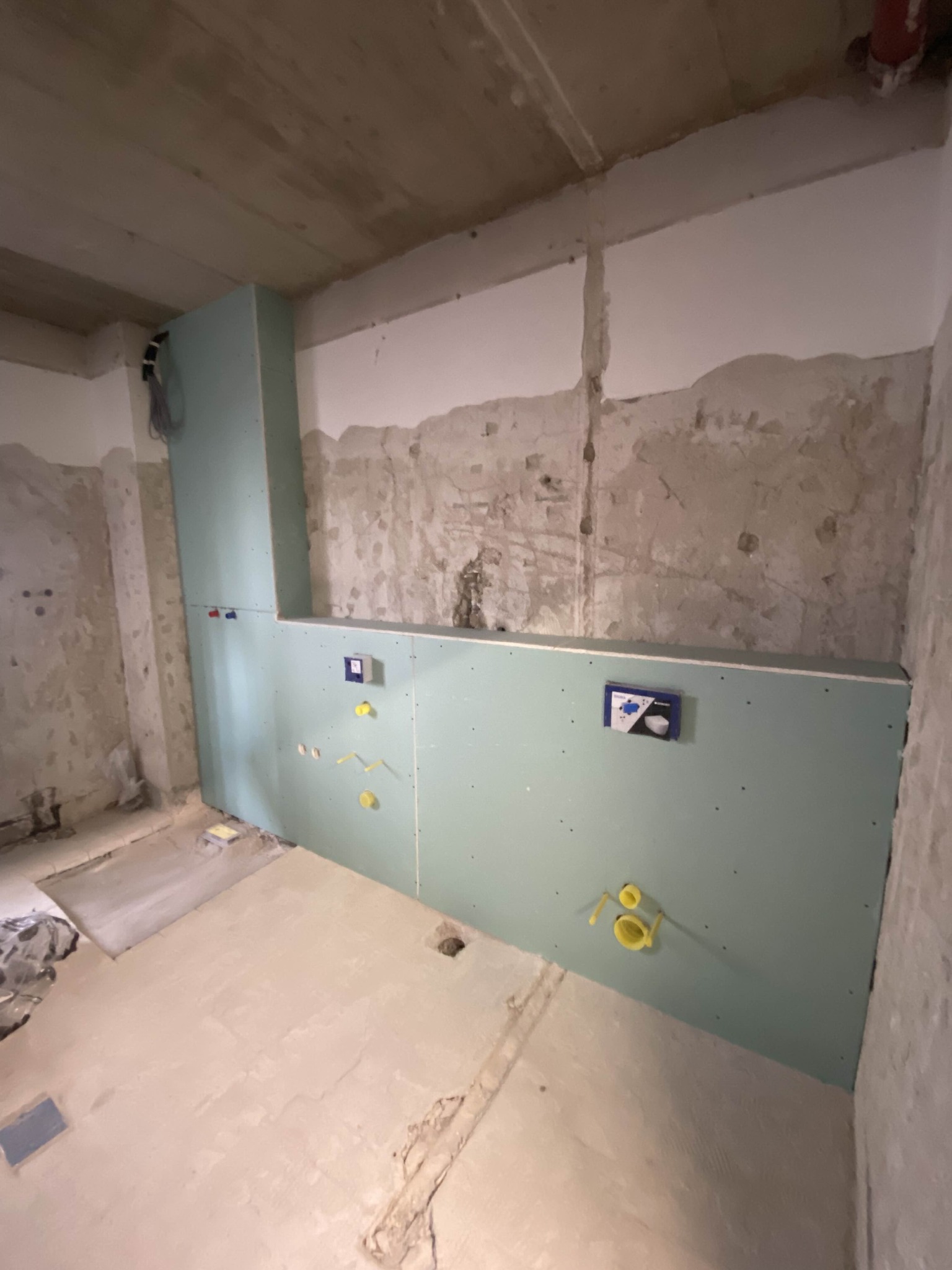 Wnętrze łazienki w trakcie remontu: ściany z surowego betonu i fragment zabudowy z płyty karton-gips z widocznymi instalacjami wodno-kanalizacyjnymi.