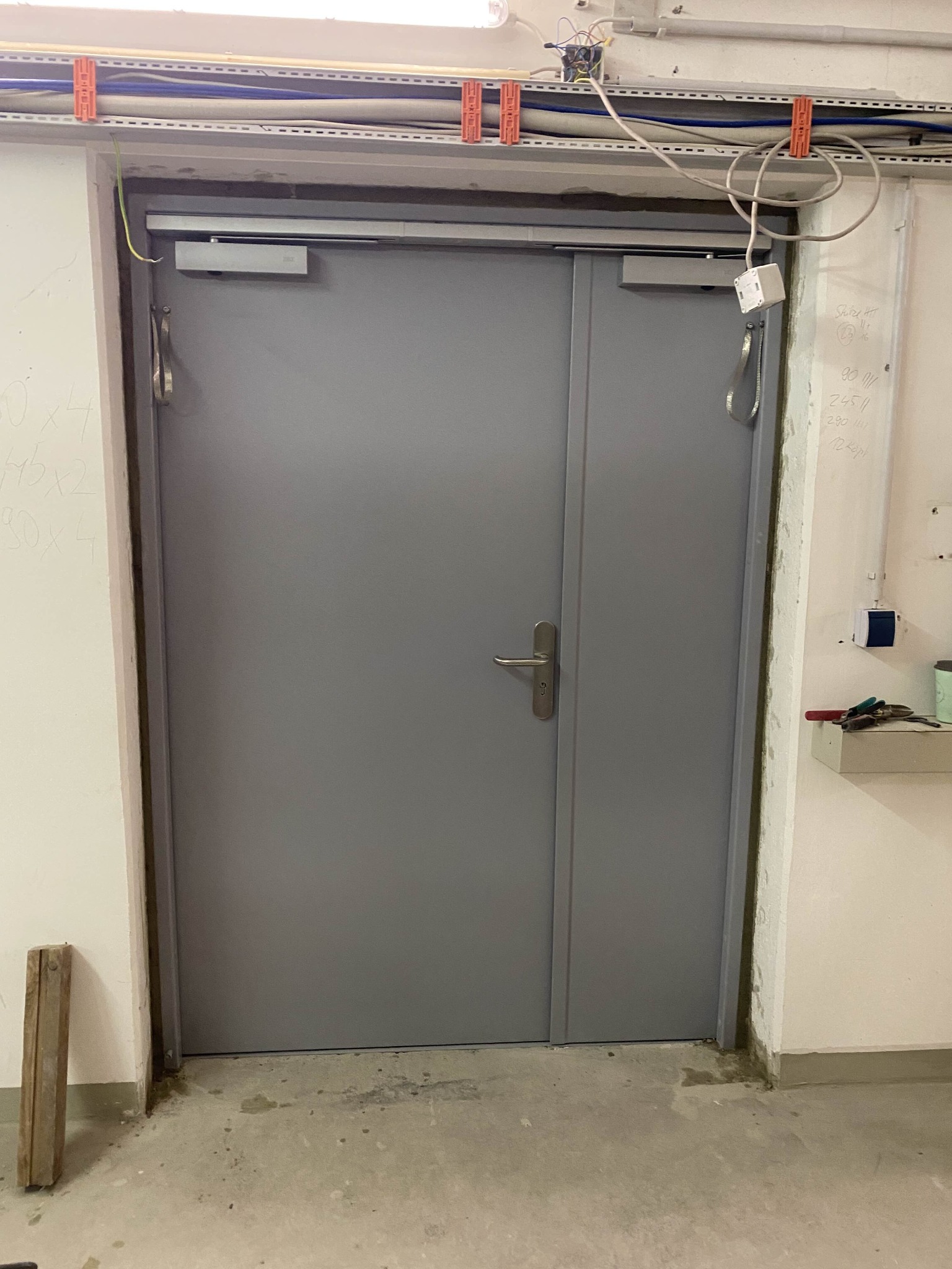 Szare, dwuskrzydłowe drzwi stalowe z samozamykaczami w surowym wnętrzu. Widoczne instalacje pod sufitem i ślady po pracach wykończeniowych na ścianach.