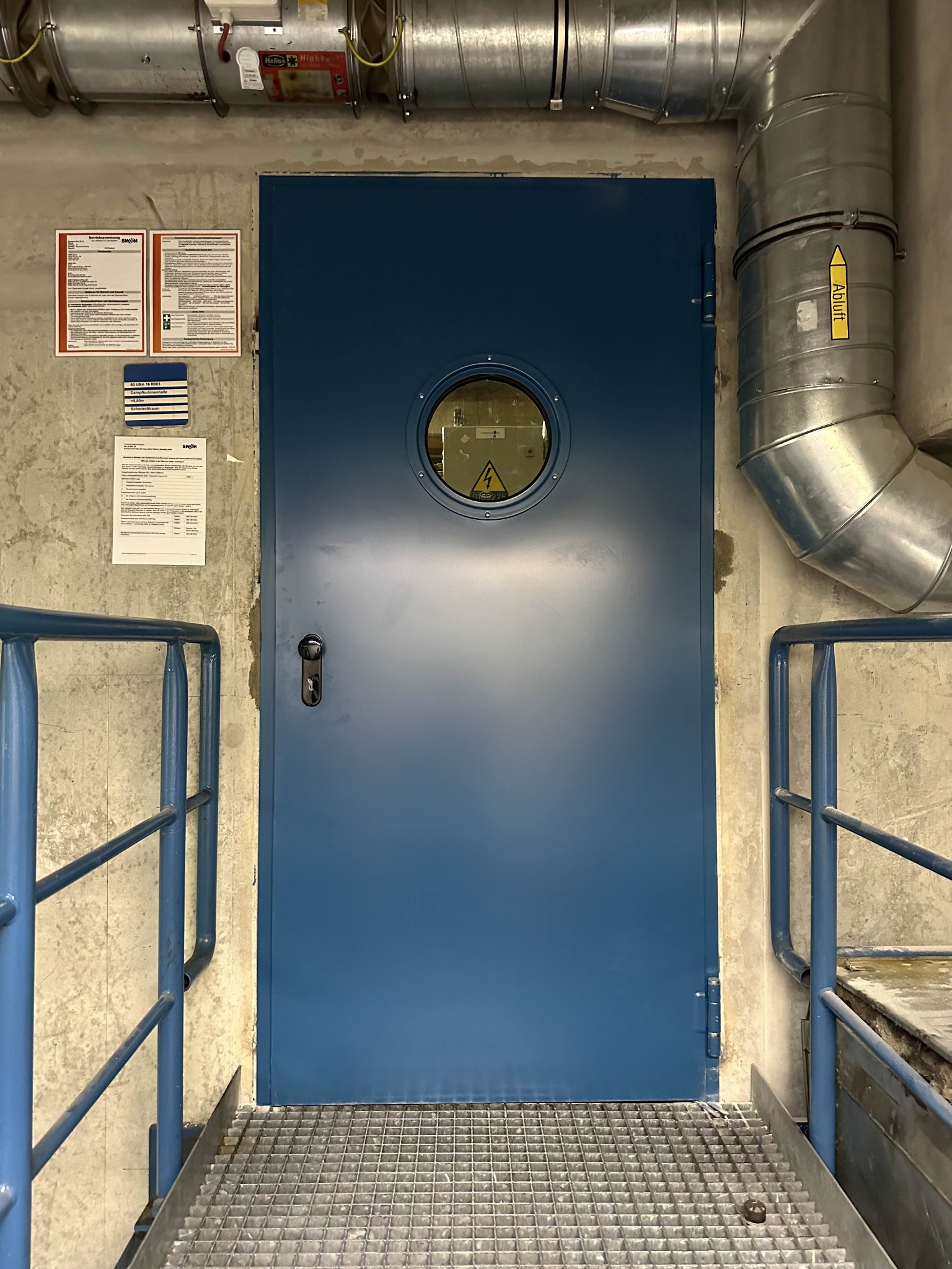 Niebieskie, metalowe drzwi techniczne z okrągłym okienkiem, widoczne na tle betonowej ściany z instalacjami wentylacyjnymi i schodami z metalowej kraty.