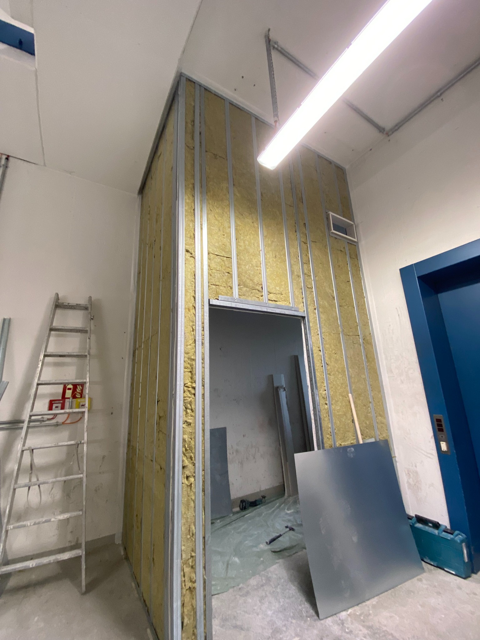 Montaż ścianki działowej z izolacją wełną mineralną, widoczne profile stalowe, drabina i narzędzia. Etap prac wykończeniowych w pomieszczeniu z niebieskimi drzwiami.