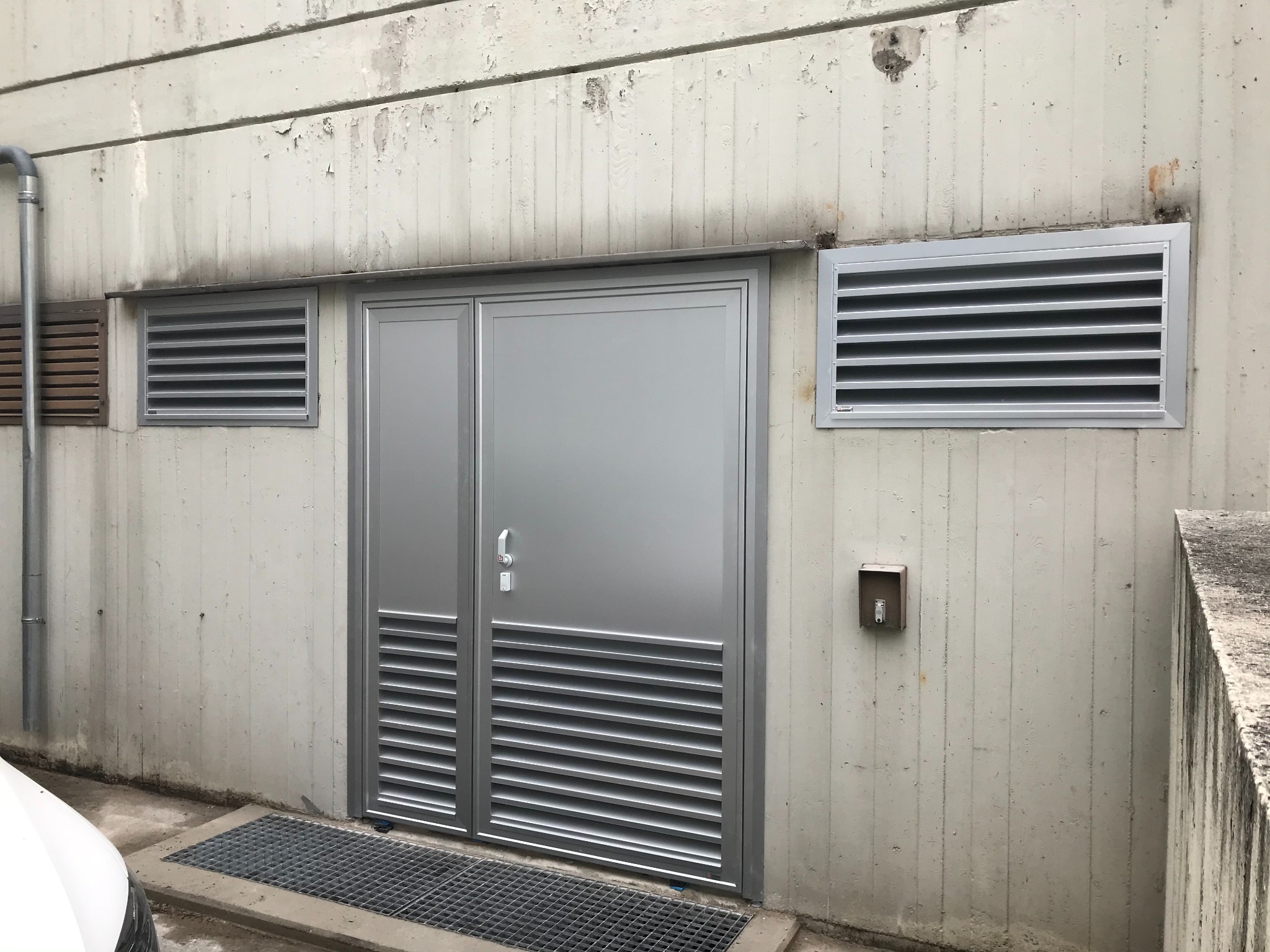 Szare, metalowe drzwi z kratkami wentylacyjnymi w betonowej ścianie. Widoczny fragment kratki ściekowej i rury spustowej. Surowy, industrialny wygląd.