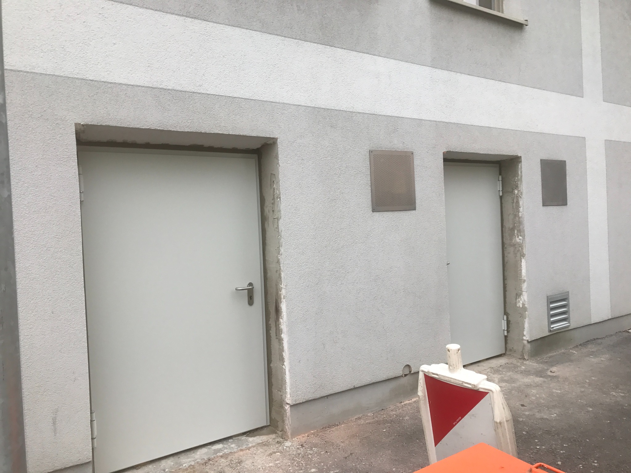 Dwa białe, metalowe drzwi zewnętrzne w surowym otoczeniu, z widocznymi śladami prac wykończeniowych na ścianie. Na ścianie kratki wentylacyjne i fragment bariery ostrzegawczej.