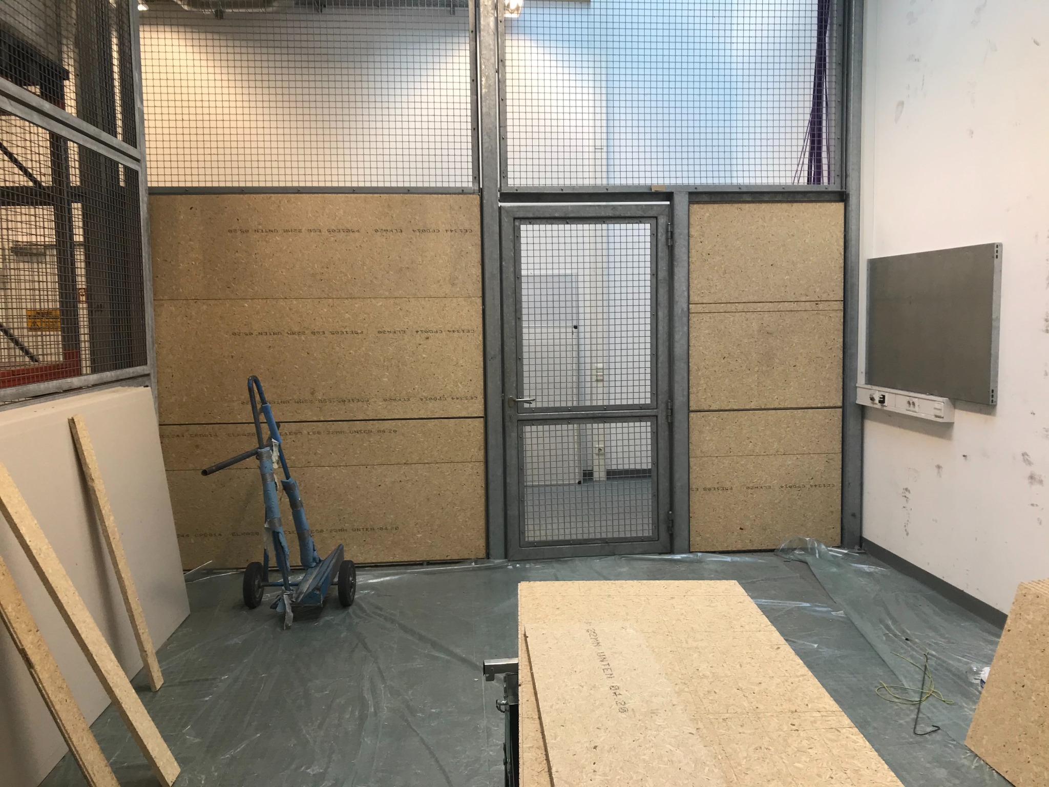 Montaż płyt OSB na metalowej konstrukcji z siatką, tworzących ścianę z drzwiami. W tle wózek transportowy i płyty. Podłoga zabezpieczona folią.