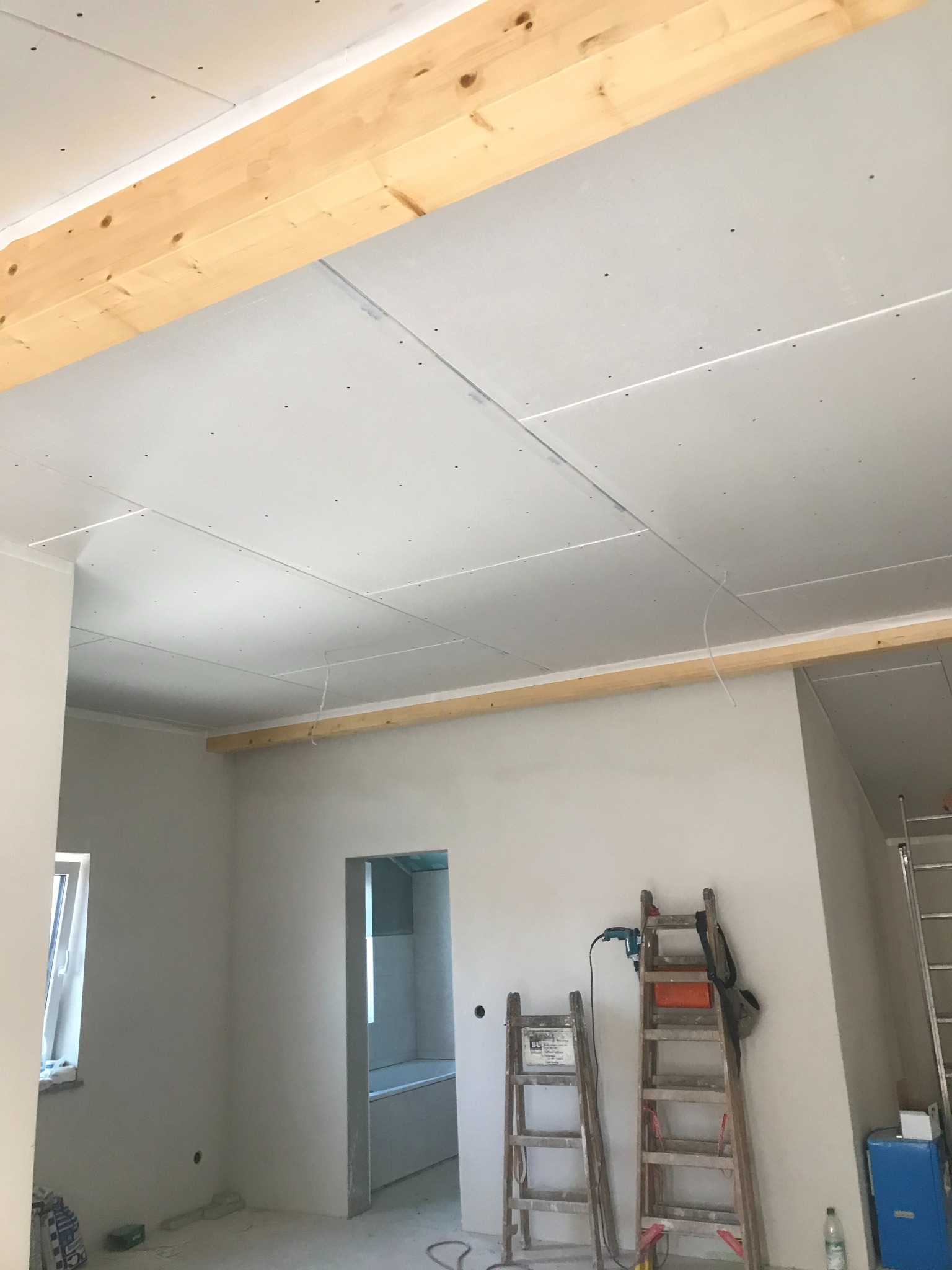 Wnętrze w trakcie montażu płyt karton-gips na suficie, widoczne drewniane belki, drabiny i narzędzia. Prace wykończeniowe w jasnym pomieszczeniu z oknem i otwartym przejściem.